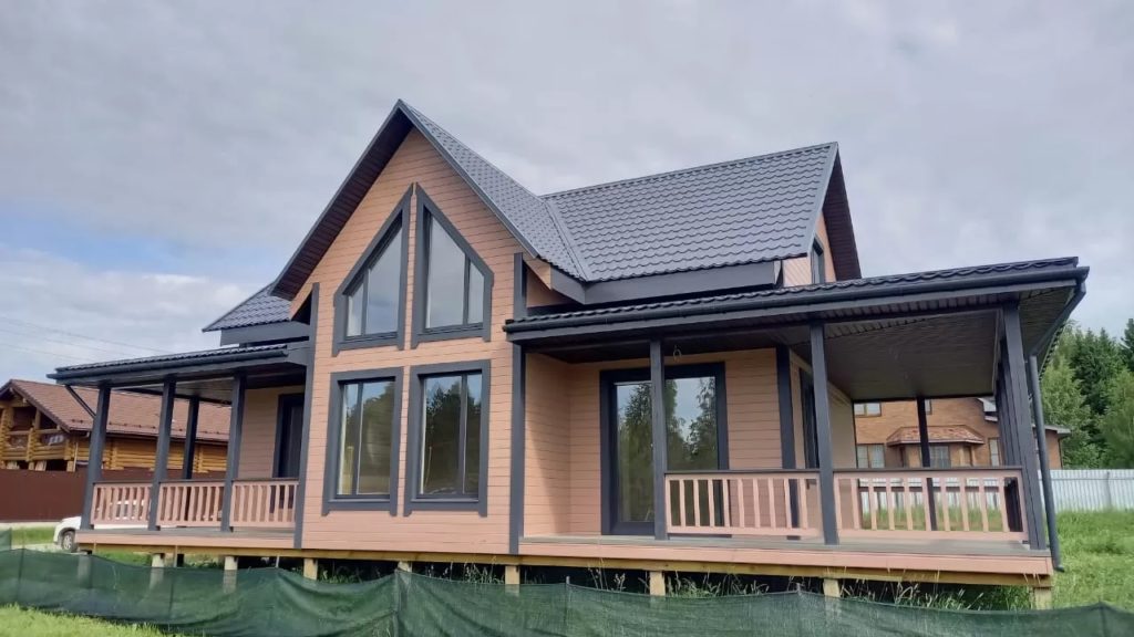 Современный деревянный дом, построенный в стиле дома из клееного бруса, с большими окнами и темно-серой крышей стоит на травянистом участке. Небо пасмурное, на заднем плане видны соседние дома и деревья.